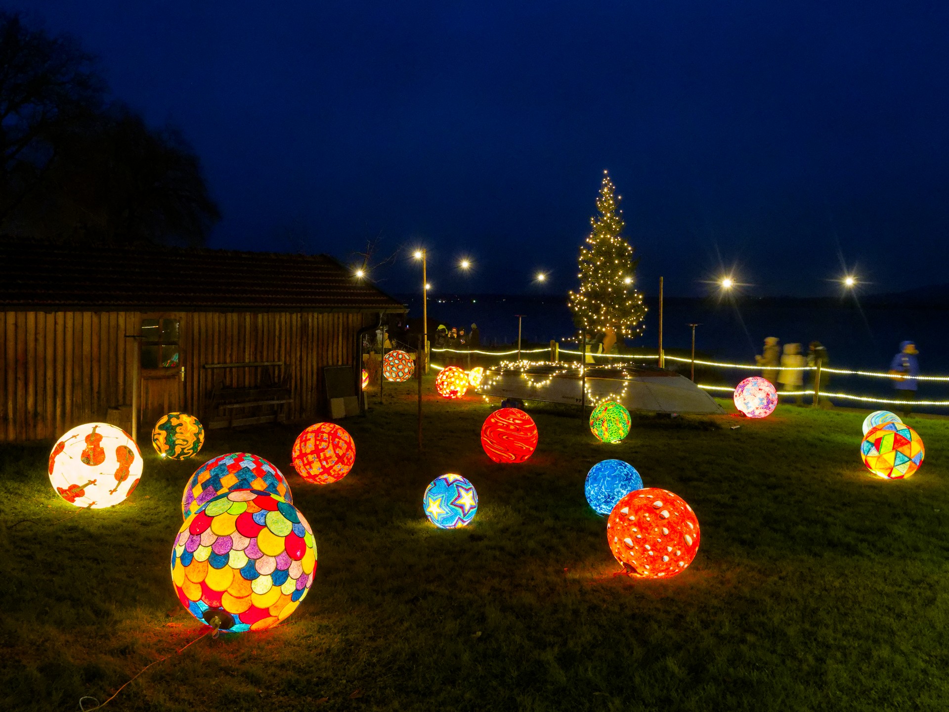 Große bunt bemalte Kugeln liegen von innen beleuchet in Dunkelheit einer Wiese. Im Hintergrund sieht man einen beleuchteten Weihnachtsbaum.