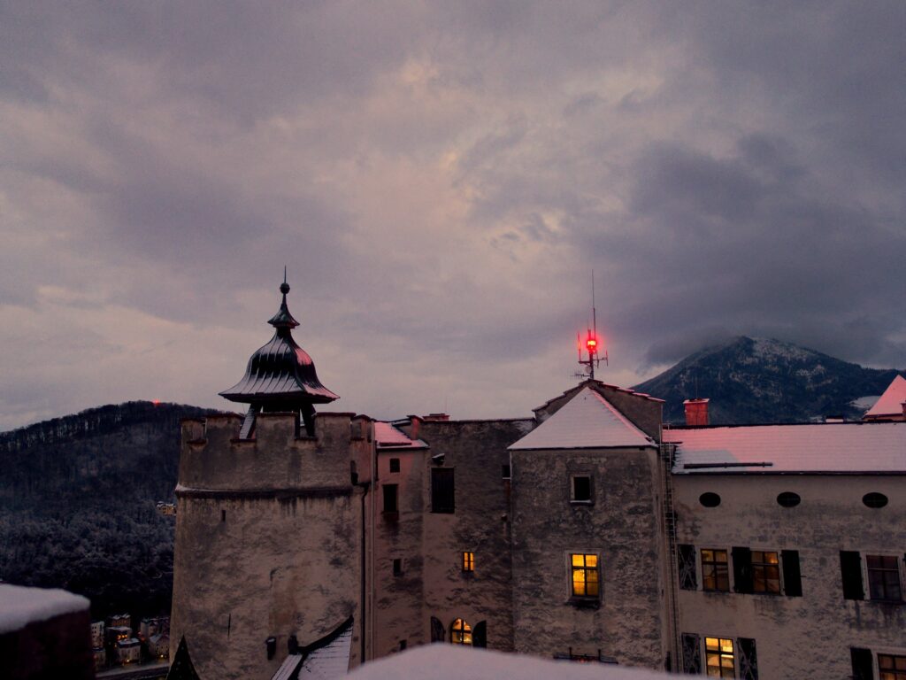 Blick auf den leicht verschneiten Glockenturm der Festung Salzburg in der Abenddämmerung mit roten Positonslichtern auf dem Kapuzinerberg und Gaisberg im Hintergrund.